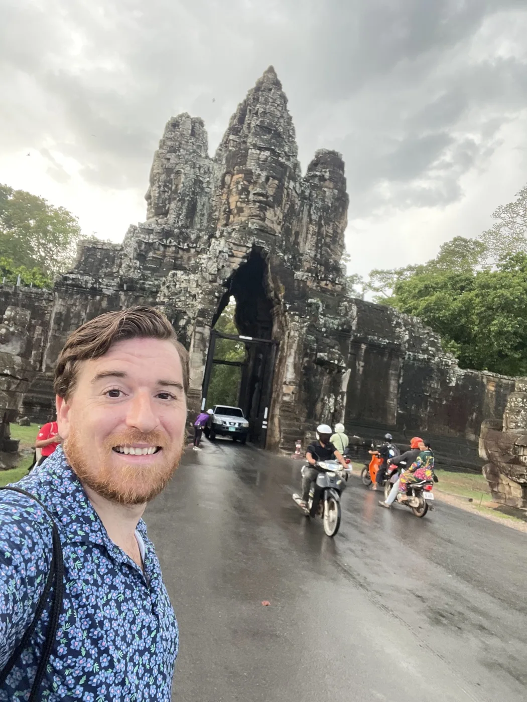 Jim posing in front of Bayon Temple stone towers with motorcyclists passing through