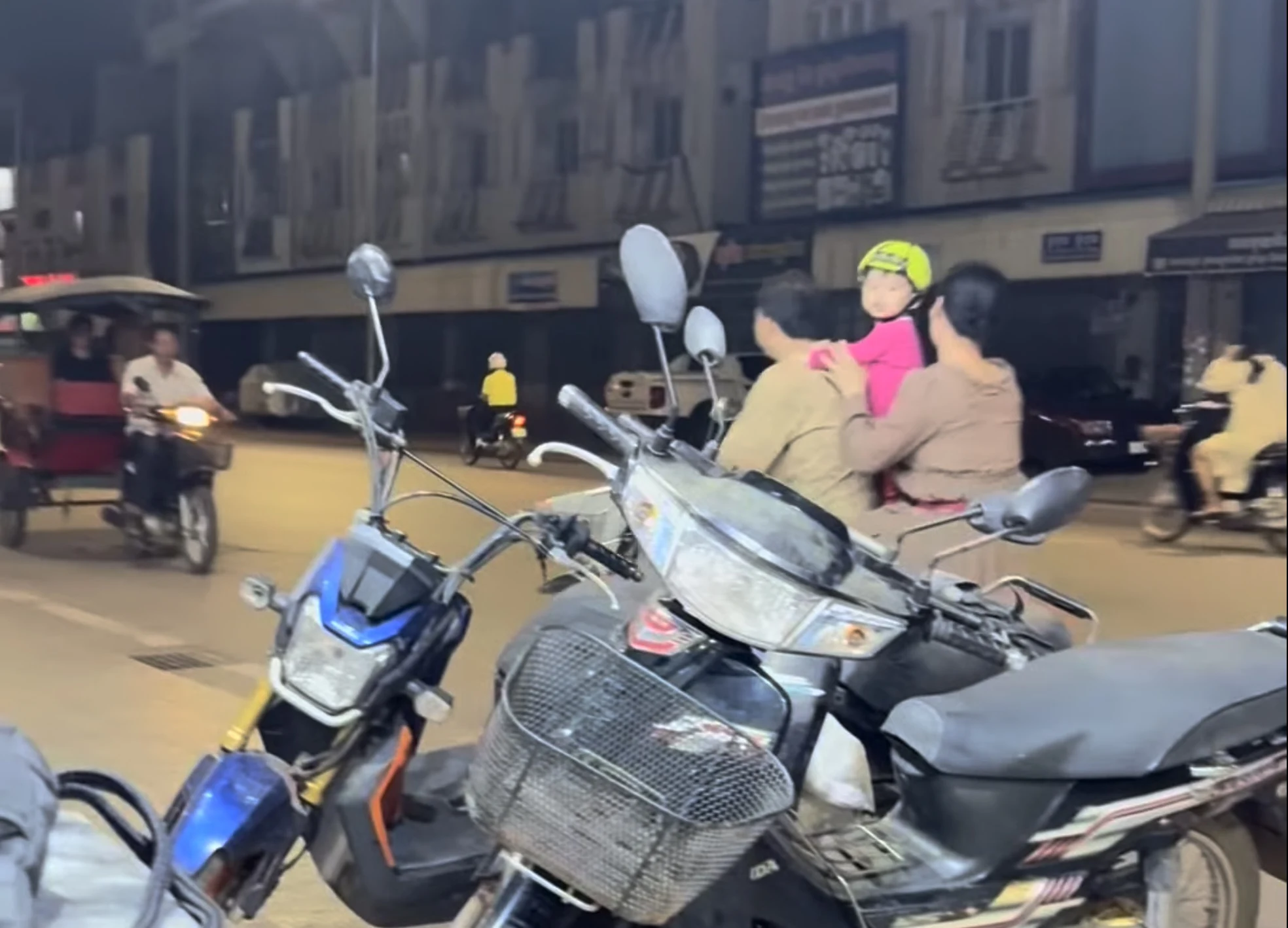 A small child wearing a bright helmet stands out amid a sea of helmetless motorcycle riders in Cambodian night traffic