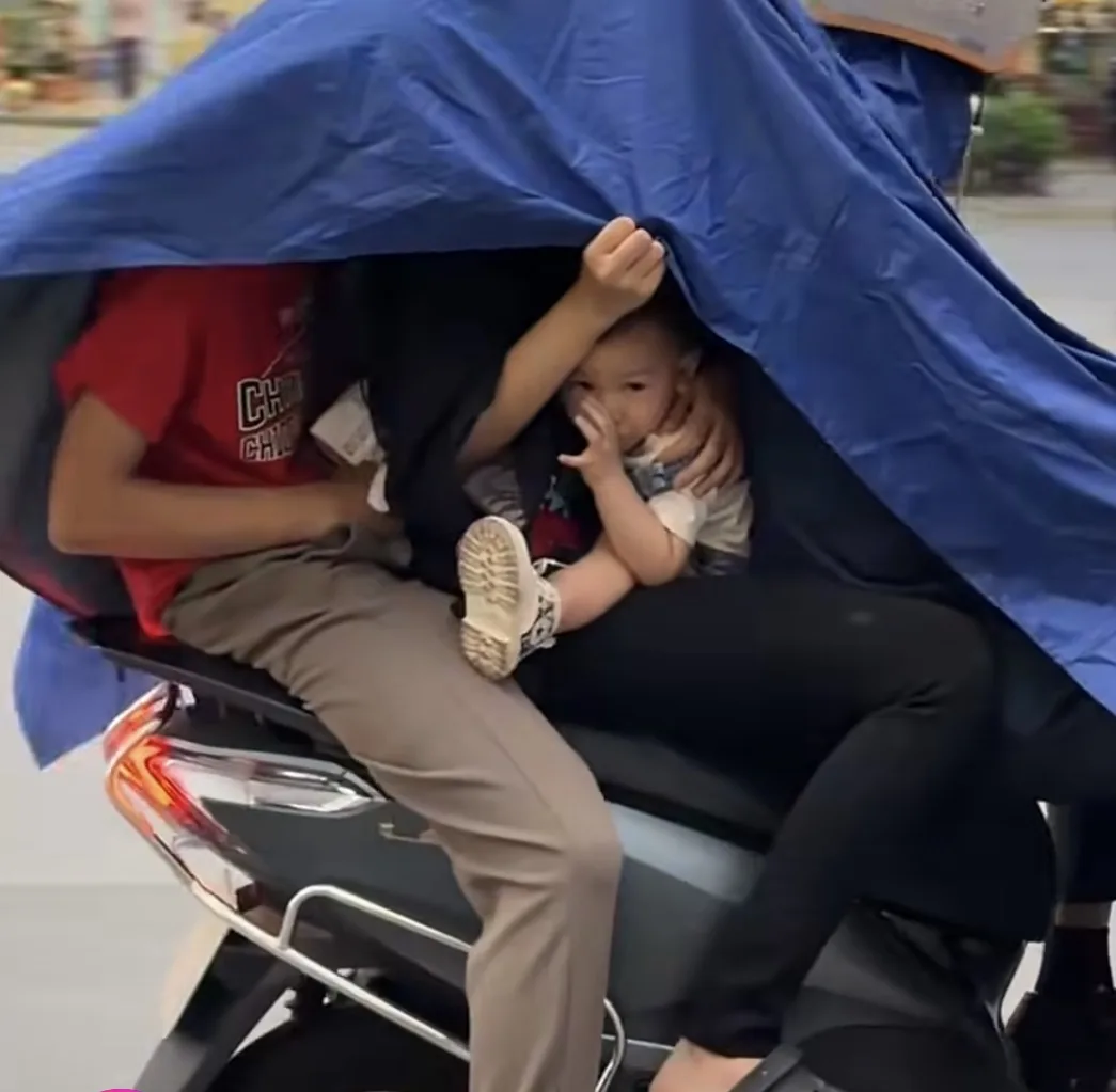 Young child riding on a motorcycle without a helmet beneath a rain cover