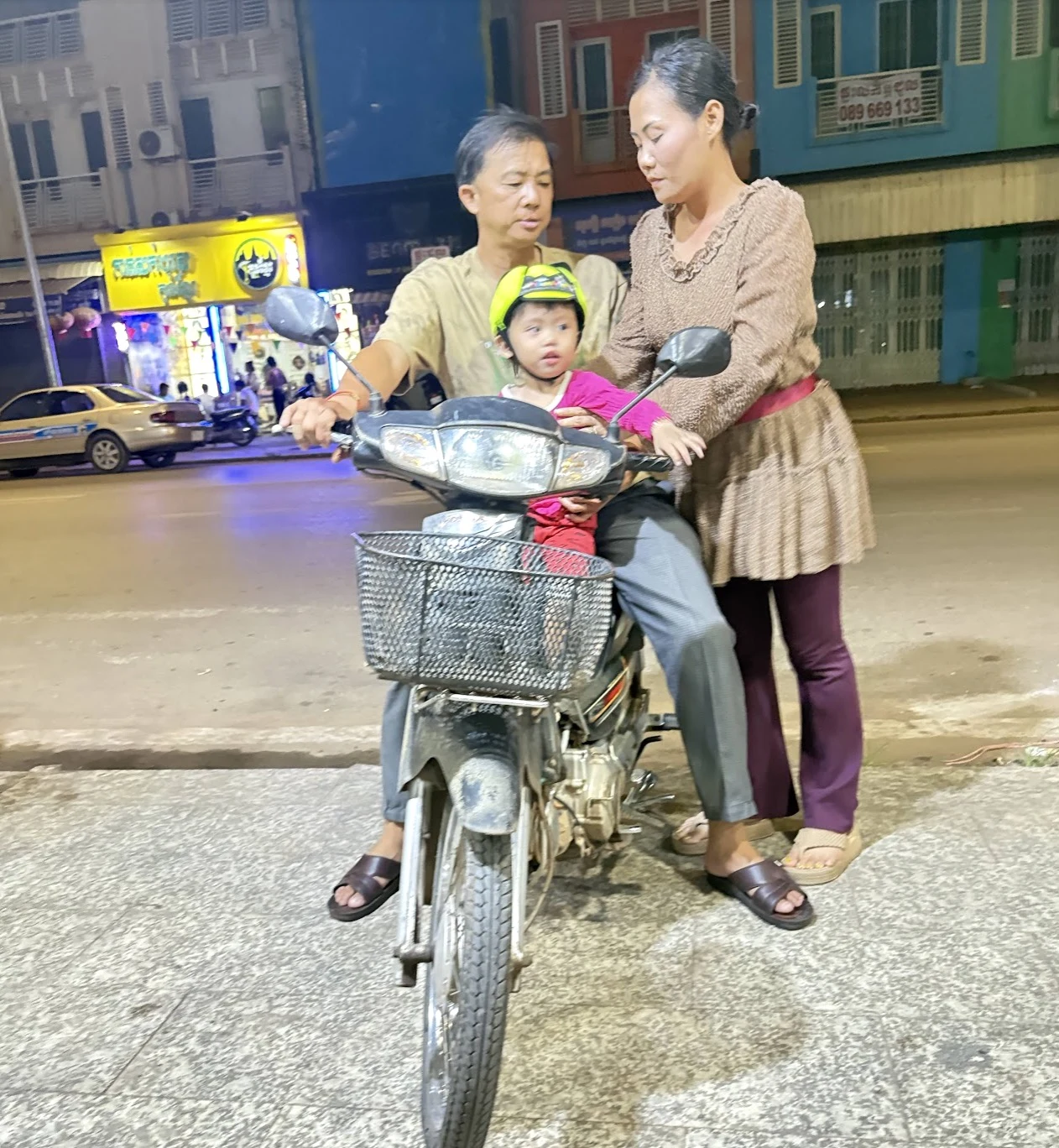 Young child in a yellow helmet seated between parents on a motorcycle at night — the only protected person on the road