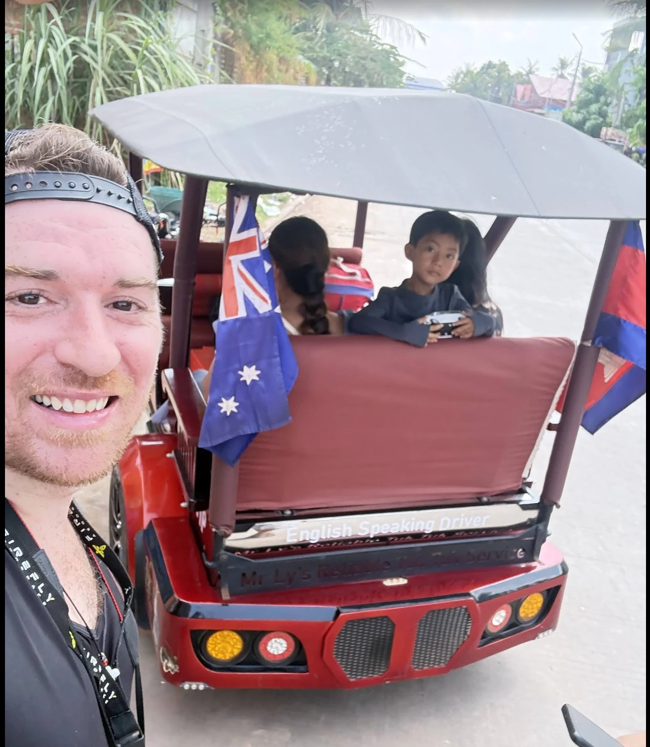Jim smiling with two Cambodian children riding in a tuk-tuk behind him