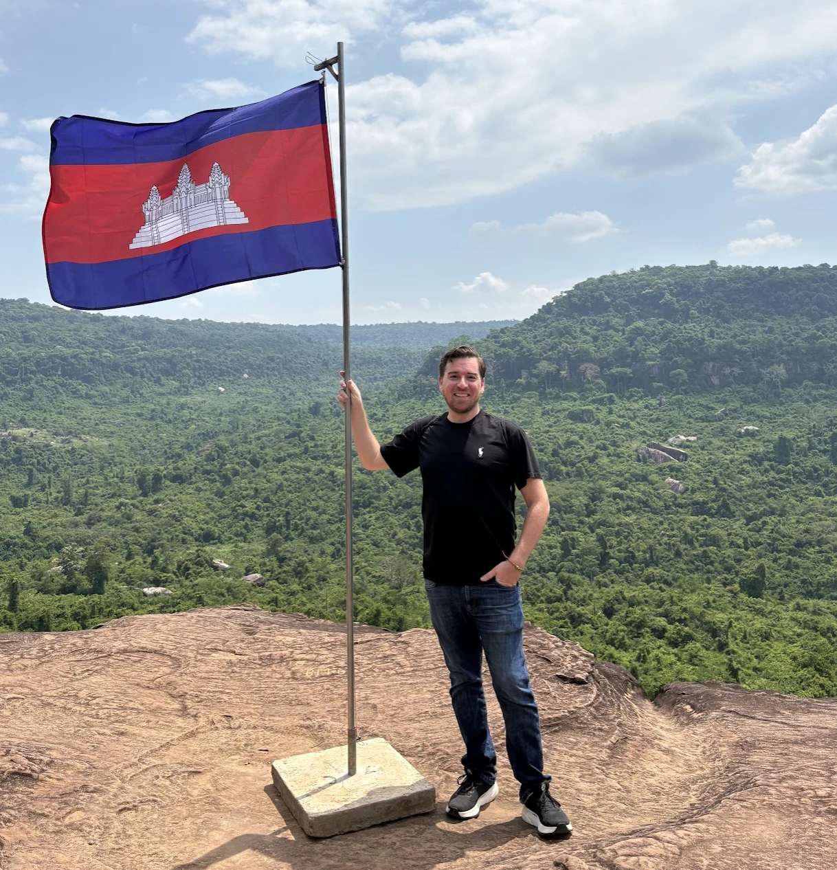 Jim standing alone on a rocky hilltop holding the Cambodian flag with forested hills stretching behind him