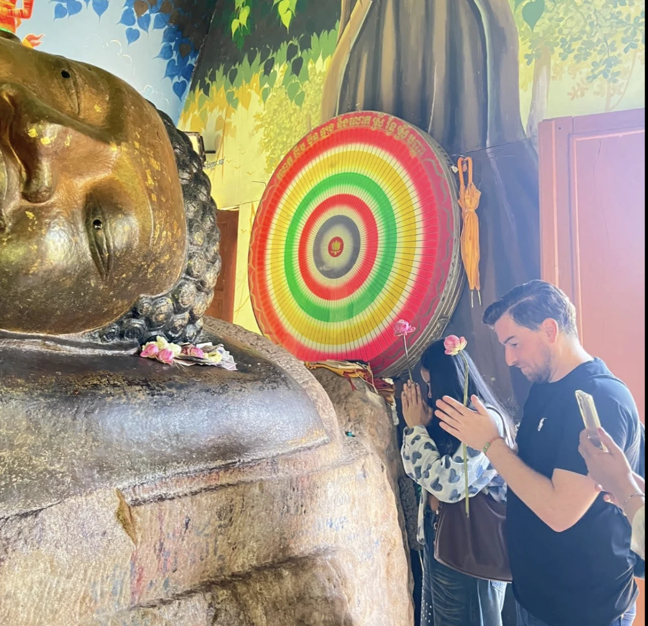 Jim praying with hands clasped before a large golden reclining Buddha statue inside a Cambodian temple
